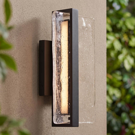 A modern home exterior with a textured black Kendrick LED Outdoor Wall Light with seeded glass glowing against a beige stucco wall at twilight near leafy foliage.