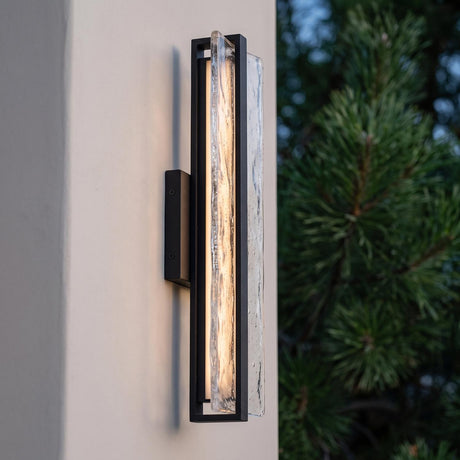A modern home exterior with a Kendrick LED Outdoor Wall Light in textured black mounted on a white stucco pillar beside a pine tree at dusk.