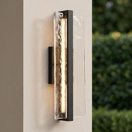 A modern patio setting with a Kendrick LED Outdoor Wall Light in textured black and seeded cast glass glowing on a beige stucco pillar beside green garden hedges.