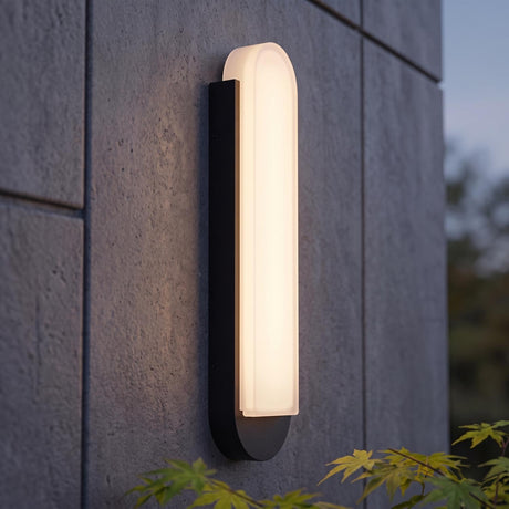 A modern home exterior with a textured black Locke Outdoor LED Wall Sconce glowing on a gray concrete panel wall at dusk, framed by green foliage.