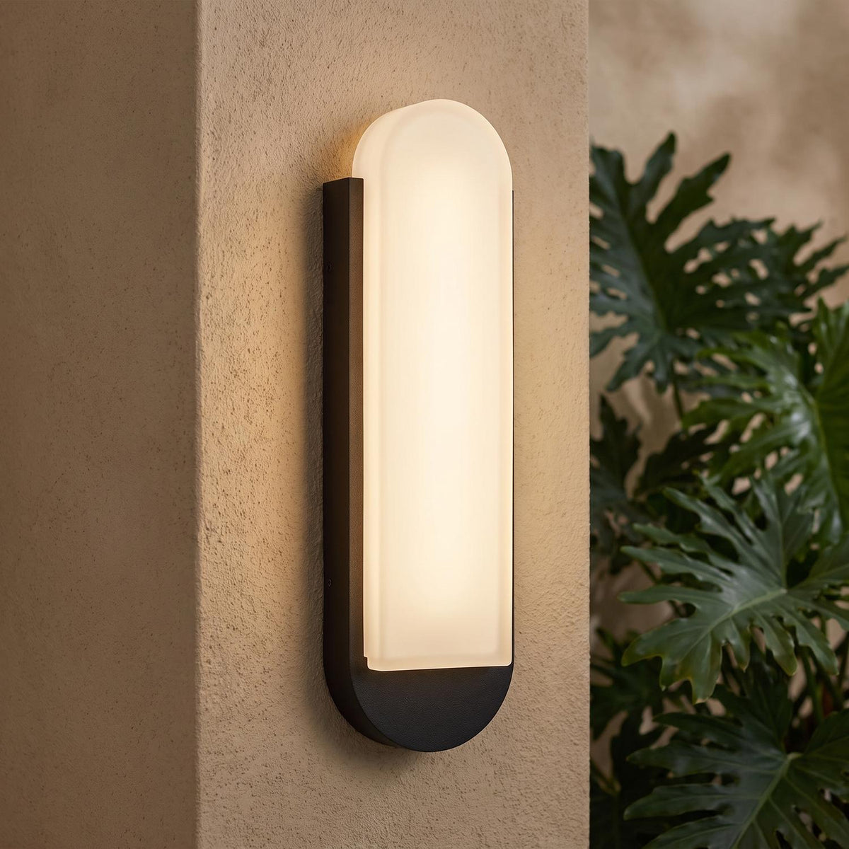 A Mediterranean home exterior with the Locke Outdoor LED Wall Sconce in textured black and frosted glass glowing on a stucco pillar at dusk near large tropical leaves.