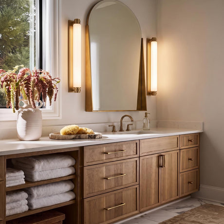 A contemporary bathroom with two Lavinia LED wall sconces in aged brass and opal glass flanking an arched mirror above a light wood vanity and white marble countertops.