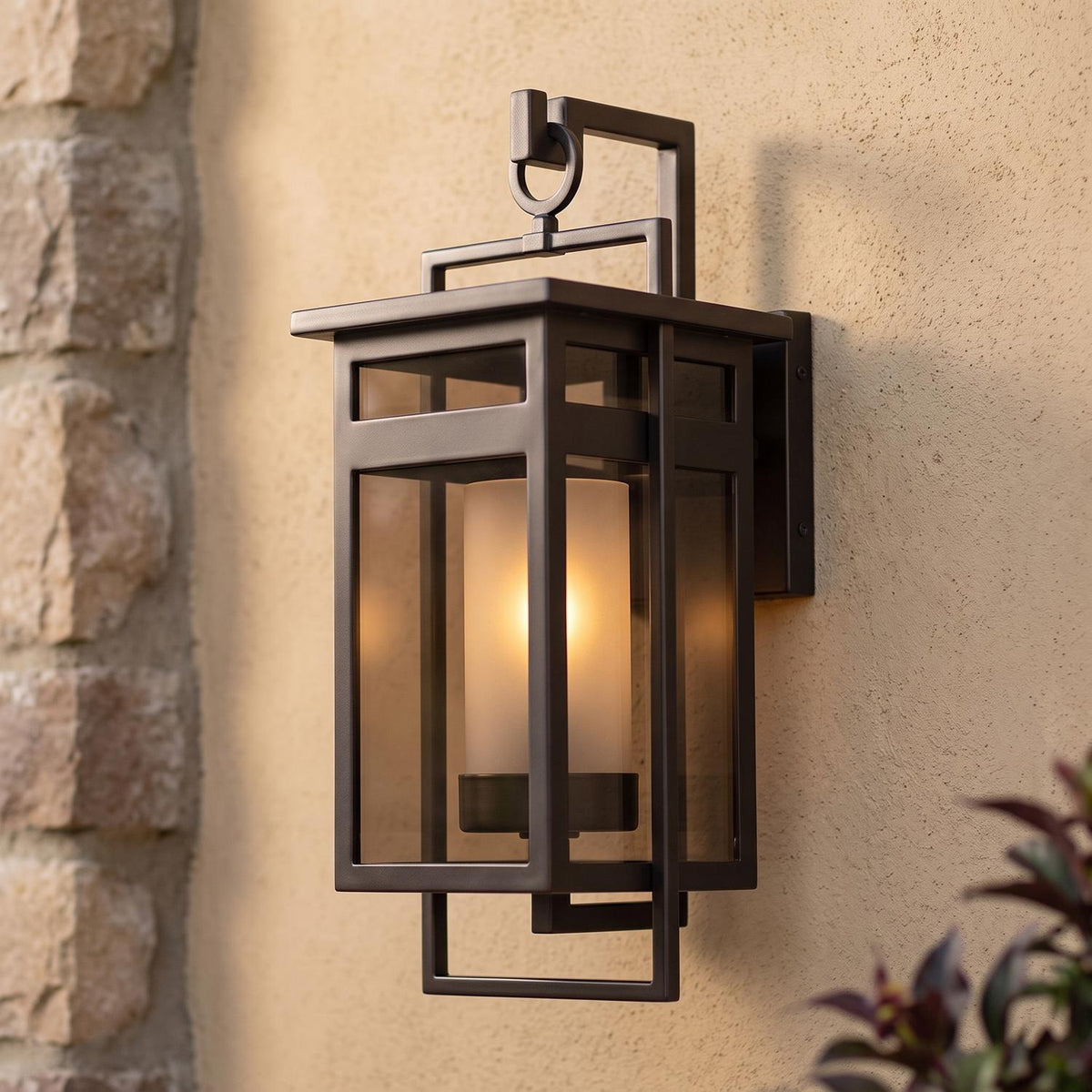A Mediterranean home exterior with the Lansing Outdoor Wall Light in dark bronze glowing on a tan stucco wall at dusk, beside a textured stone pillar.