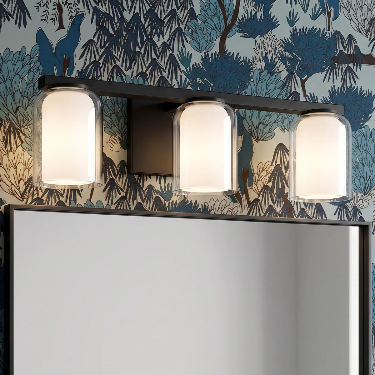 A contemporary bathroom with the Orlando 3 Light Vanity 23", Matte Black featuring clear and frosted glass shades above a black framed mirror against botanical wallpaper.