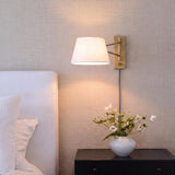 A transitional bedroom with a Barton Medium Swing Arm Wall Sconce in aged brass and white linen mounted beside a white headboard and black nightstand.