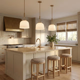 A transitional kitchen with three Stratford Small Pendants in aged brass and opal glass hanging over a beige stone island with neutral cream shaker cabinets.