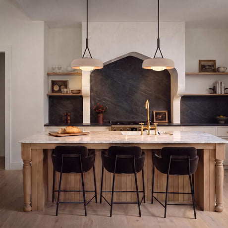 A modern farmhouse kitchen featuring two Stratford Medium Pendants in aged iron and beige over a white marble island with wood cabinets and dark stone backsplash.