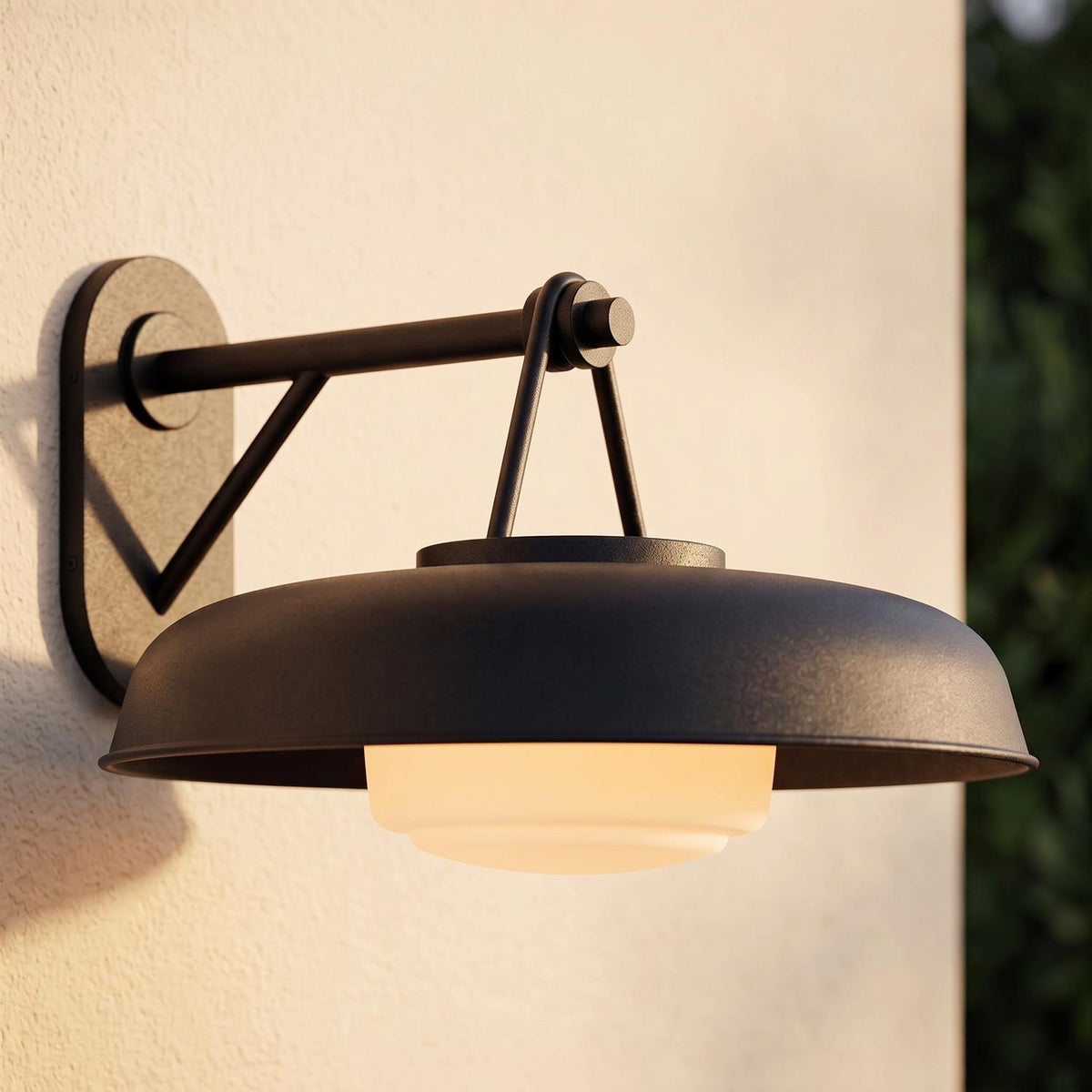 A modern home exterior with the Stratford Outdoor Wall Sconce, Medium, Textured Black glowing on a beige stucco wall at dusk beside dark green foliage.