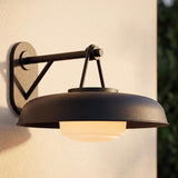 A modern home exterior with the Stratford Outdoor Wall Sconce, Medium, Textured Black glowing on a beige stucco wall at dusk beside dark green foliage.