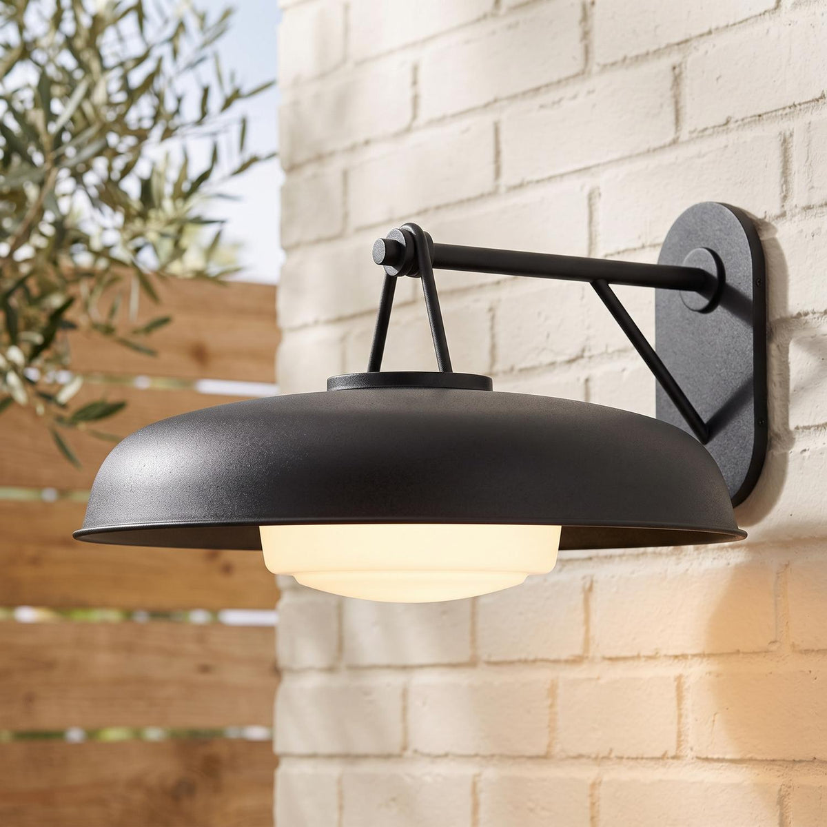 A modern farmhouse patio featuring the Stratford Outdoor Wall Sconce, Large, Textured Black mounted on a white brick wall beside an olive tree and wooden fence.