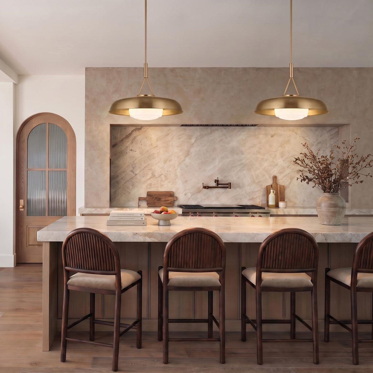 A transitional kitchen featuring two Stratford Large Pendants in aged brass suspended over a white marble island with dark wood bar stools and a stone backsplash.