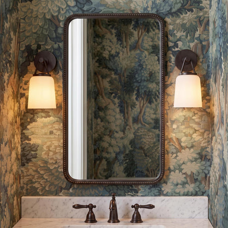 A traditional bathroom with two aged iron and opal glass wall sconces flanking a beaded mirror above a white marble vanity and forest landscape wallpaper.