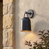 A Mediterranean home exterior with a Stratford Outdoor Wall Sconce, Small, Textured Black glowing on a beige stucco wall beside a stone pillar with dark green foliage.
