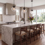 A transitional kitchen with three matte black Whitaker Pendants with clear ribbed glass suspended over a white marble waterfall island featuring greige cabinets and wood barstools.