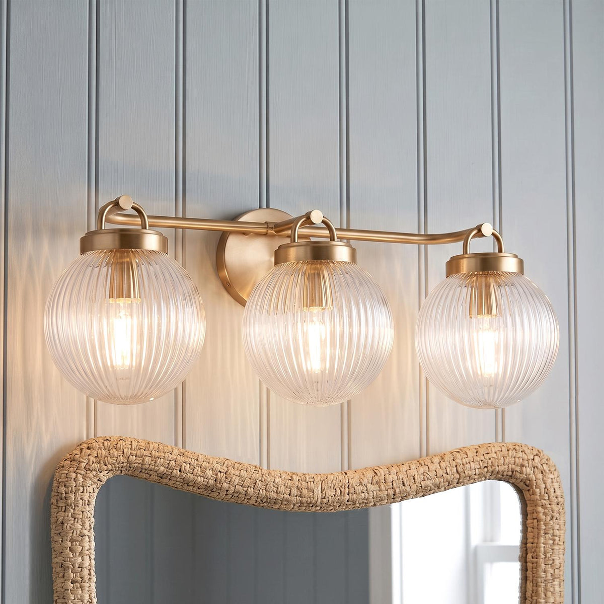 A coastal bathroom with the Whitaker 3 Light Vanity 24", Satin Brass and Clear Ribbed Glass installed over a woven rattan mirror and light blue shiplap walls.