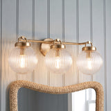 A coastal bathroom with the Whitaker 3 Light Vanity 24", Satin Brass and Clear Ribbed Glass installed over a woven rattan mirror and light blue shiplap walls.