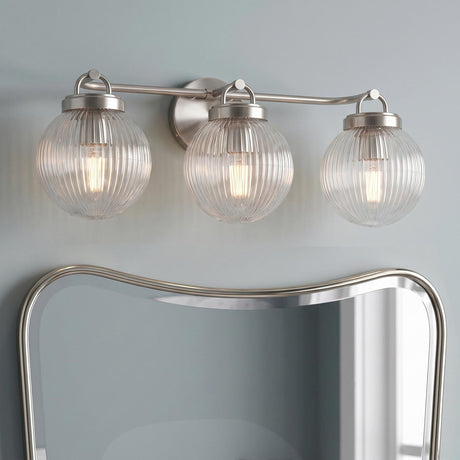 A transitional bathroom features a Whitaker 3 Light Vanity 24", Brushed Nickel and Clear Ribbed Glass fixture mounted on a light blue wall above a curved mirror.
