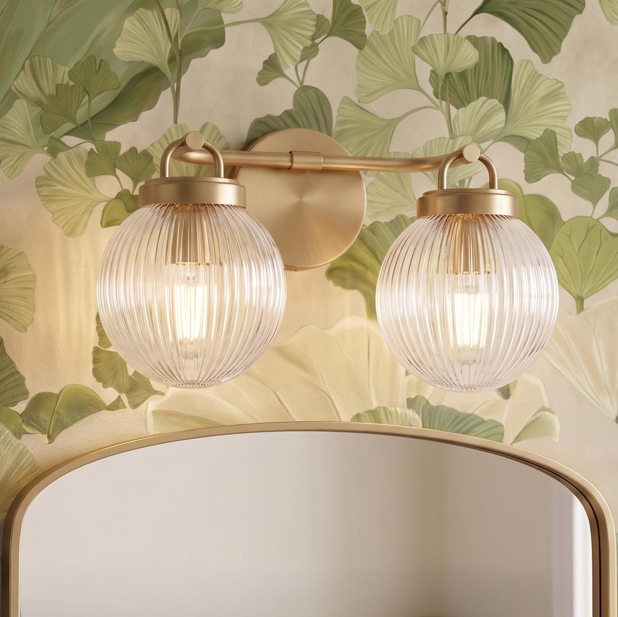 A transitional bathroom with a satin brass and clear ribbed glass vanity bar mounted above a gold arched mirror against green ginkgo leaf wallpaper.