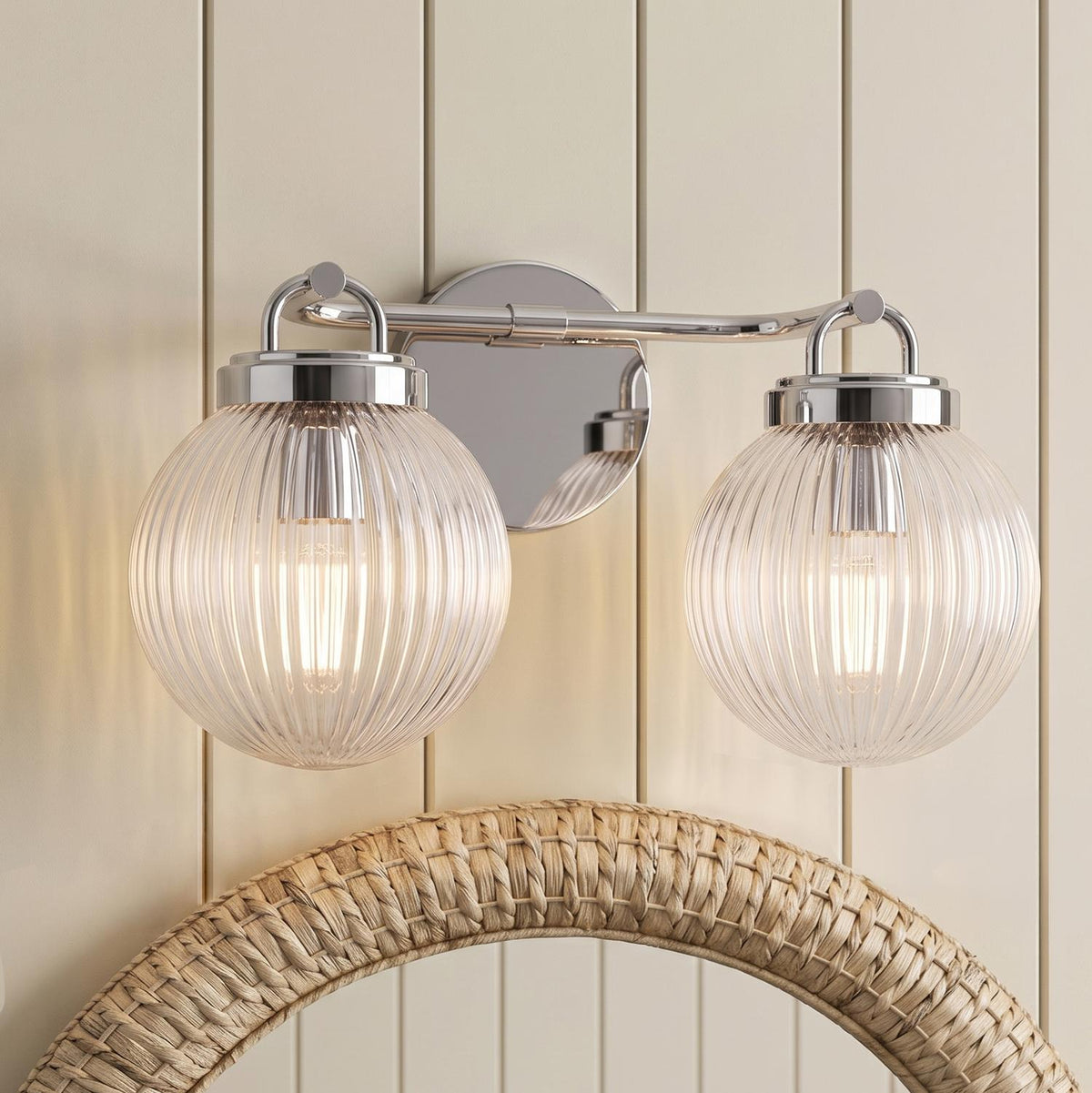 A coastal bathroom featuring the Whitaker 2 Light Vanity in chrome with clear ribbed glass globes mounted above a woven rattan mirror on cream paneled walls.
