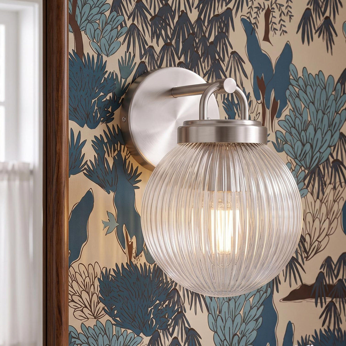 A transitional bedroom features a Whitaker Wall Sconce in brushed nickel and clear ribbed glass mounted on blue botanical wallpaper beside a dark wood mirror.