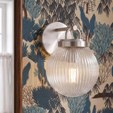 A transitional bedroom features a Whitaker Wall Sconce in brushed nickel and clear ribbed glass mounted on blue botanical wallpaper beside a dark wood mirror.