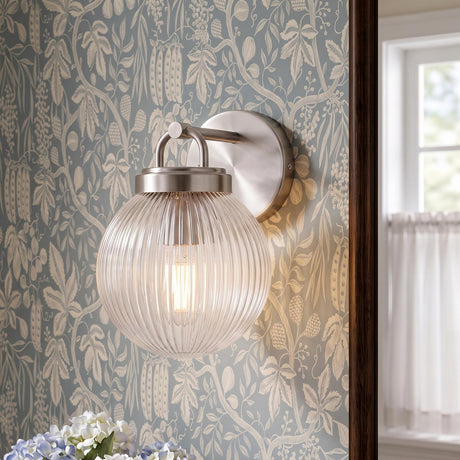 A traditional bathroom featuring a Whitaker Wall Sconce in brushed nickel and clear ribbed glass mounted on blue botanical wallpaper near a white cafe curtain.