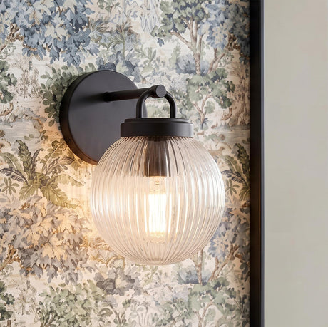 A traditional foyer featuring a matte black and clear ribbed glass Whitaker Wall Sconce 9" mounted on a wall decorated with blue and green floral wallpaper.