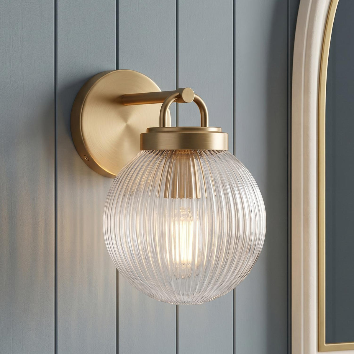 A transitional bathroom with the Whitaker Wall Sconce in satin brass and clear ribbed glass mounted on blue vertical shiplap walls beside an arched gold mirror.