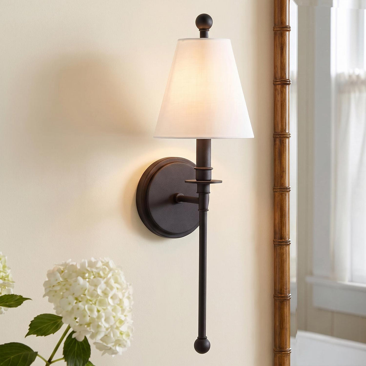 A transitional bathroom with a matte black wall sconce featuring a white linen shade mounted beside a bamboo mirror and a vase of white hydrangeas.