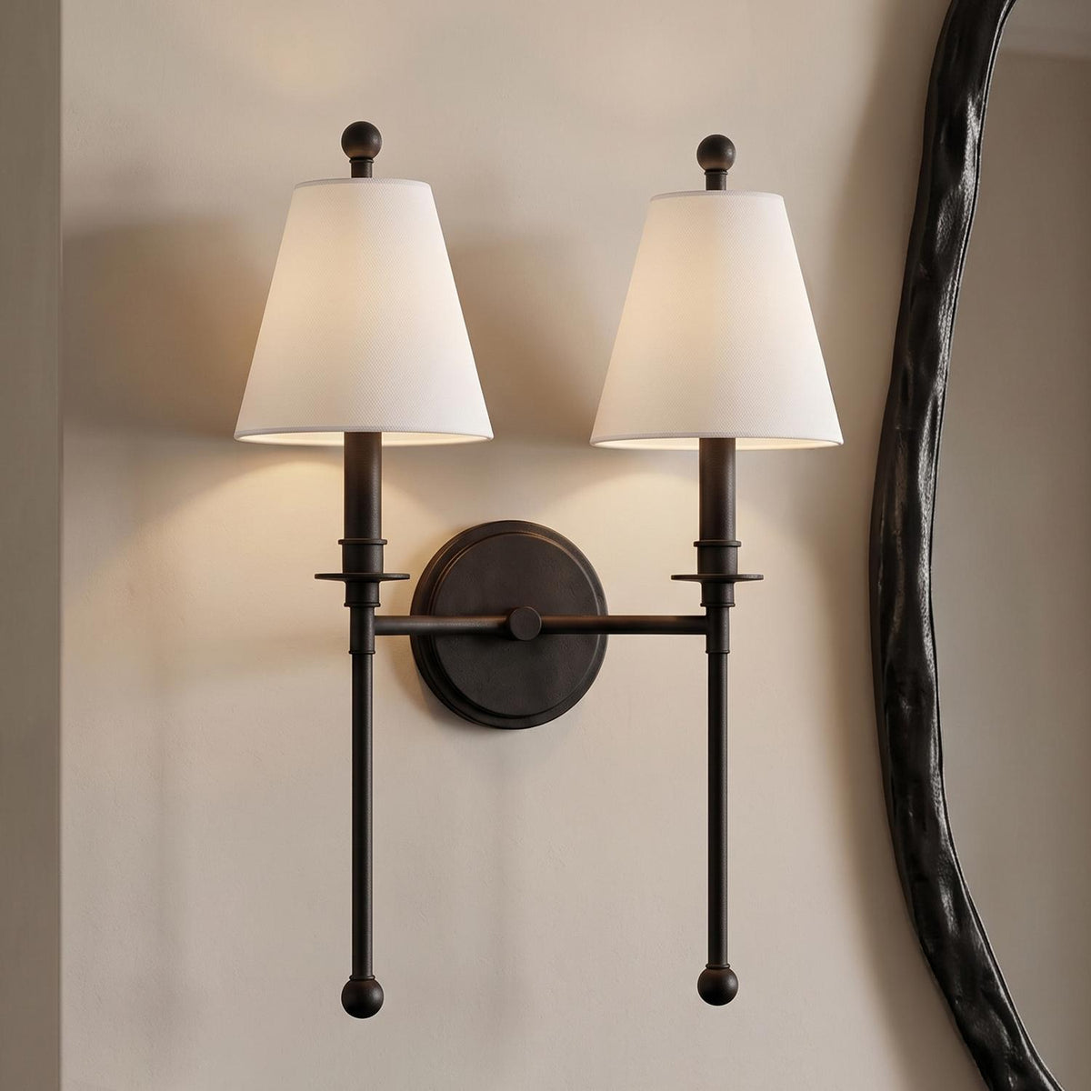 A transitional foyer featuring a forged black Bellamy double wall sconce with white fabric shades mounted on a neutral wall beside a dark textured mirror.