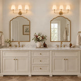 A transitional bathroom features two brushed brass 3-light vanity lights with white fabric shades mounted above arched mirrors over a marble double vanity.