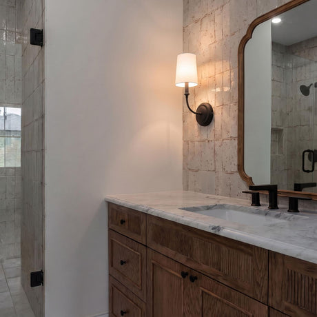 A transitional bathroom with the Estelle Wall Sconce 16", Forged Black and White Linen mounted on a beige tile wall beside a wood mirror and marble vanity.