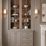 A transitional living room with Estelle Wall Sconce 16", Polished Nickel and White Linen fixtures flanking taupe glass-front cabinetry beside a carved stone fireplace.
