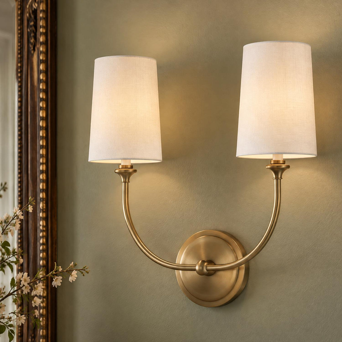 A transitional living room featuring the Estelle Double Wall Sconce 17", Brass and White Linen mounted on a muted green wall next to an ornate mirror.