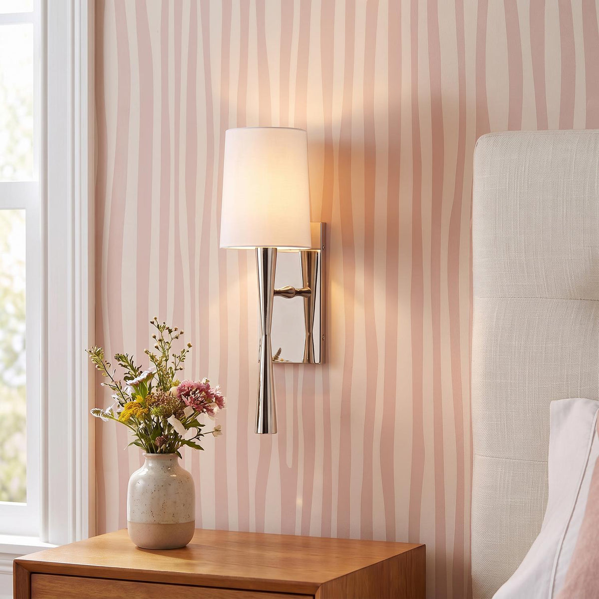 A transitional bedroom featuring the Kellan Wall Sconce 18 in Polished Nickel and White Fabric mounted on pink patterned wallpaper above a wooden nightstand.