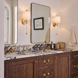 A transitional bathroom features aged brass wall sconces with white fabric shades flanking metal mirrors above a purple-veined marble countertop and dark walnut vanity.