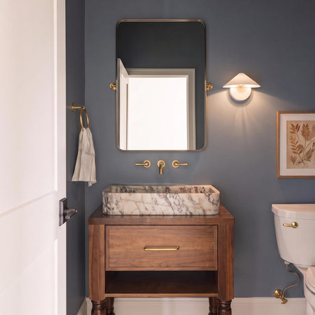 A mid-century bathroom featuring an antique white Concord Wall Sconce mounted beside a rectangular brass mirror above a marble vessel sink on a walnut vanity with blue walls.