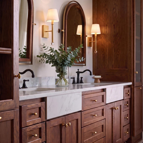 A transitional bathroom featuring two gilded brass wall sconces with fabric shades flanking arched mirrors above a marble double vanity with dark wood cabinetry.