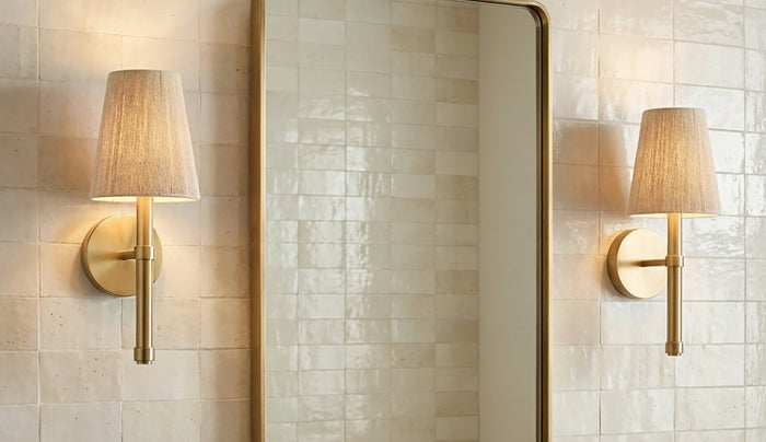 Two Evren Wall Sconces in Brass with natural Jute string shades, shown flanking a brass curved edge vanity mirror against natural stone stacked backsplash tiles.
